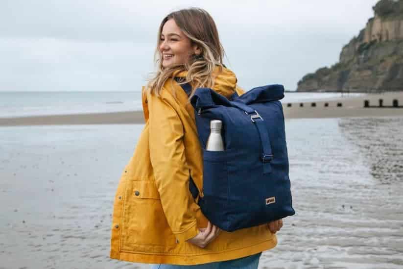 Person on a beach wearing a yellow jacket, jeans and a navy blue organic cotton backpack