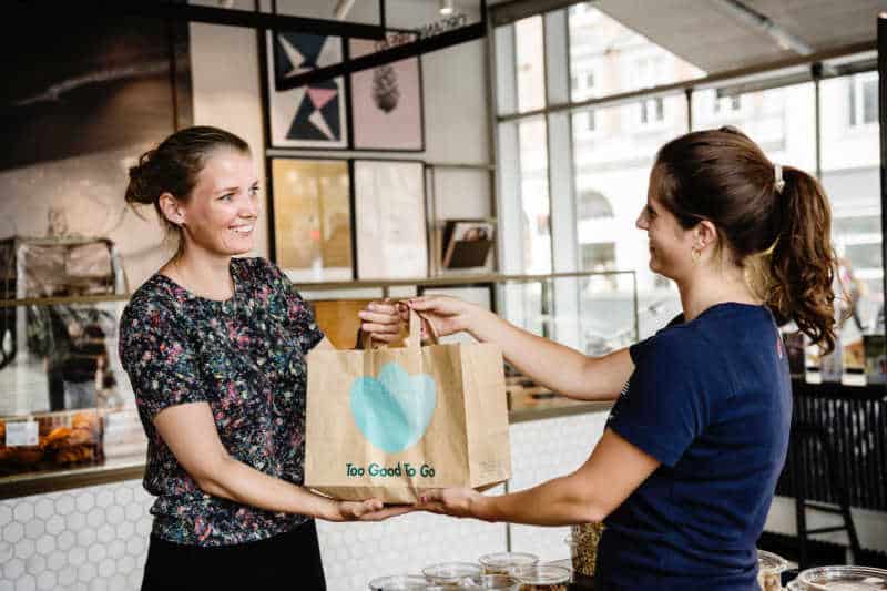 Cafe owner handing over bag off food to customer, purchased via the food waste sustainability app Too Good To Go.