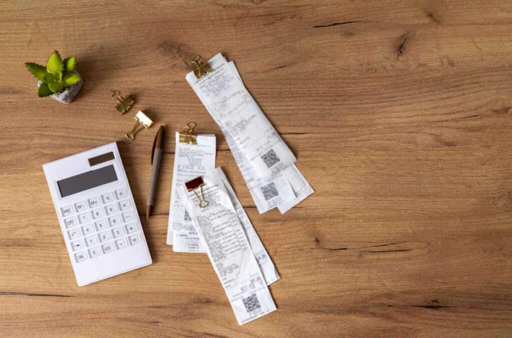 receipts on a wooden desk next to a calculator, pens, and plant.