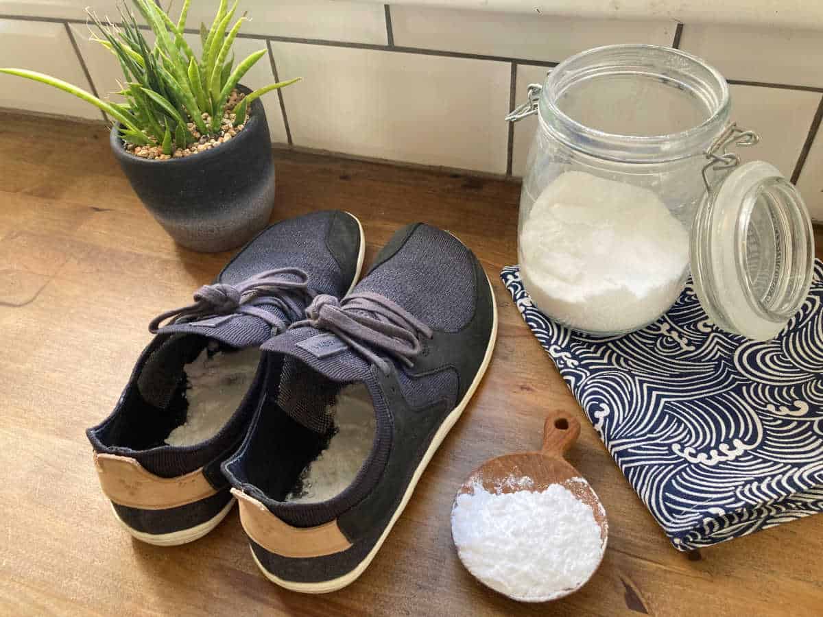 Black pair of trainers with bicarbonate of soda in them, next to  a plant, jar of bicarb and a spoon.