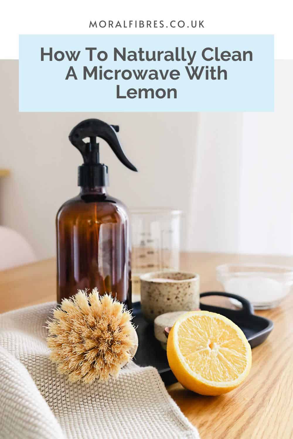 A cut lemon, amber glass spray bottle and scrubbing brush on a table, with a blue text box that reads how to naturally clean a microwave with lemon